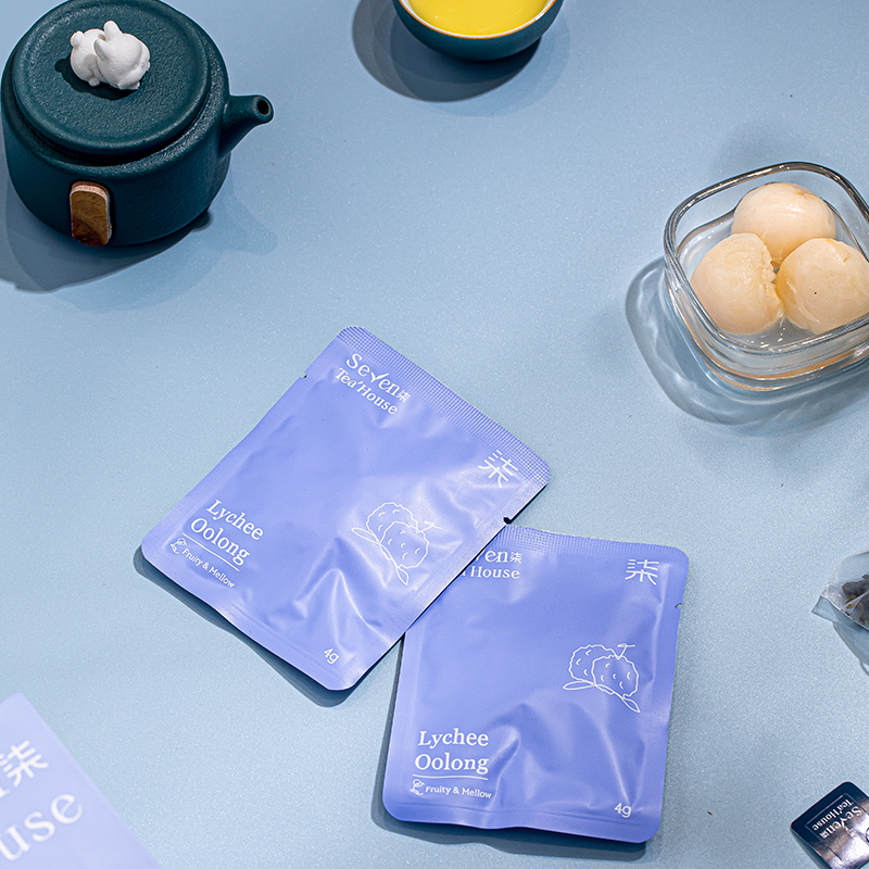 Two blue packets of lychee oolong tea bags on a blue surface surrounded by tea accessories and lychee fruit